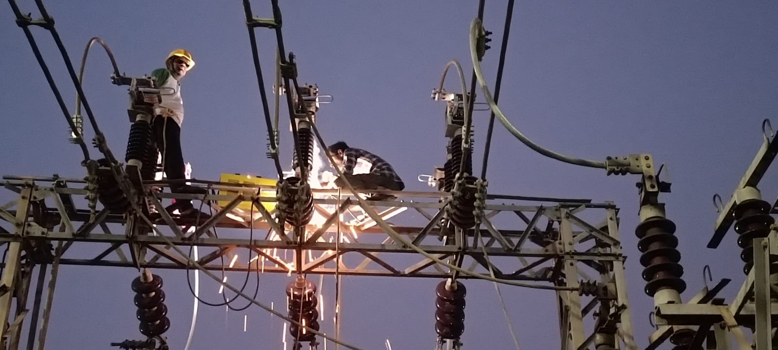 Two technicians working on a substation steel gantry structure at dusk, with sparks from cutting work near the insulators